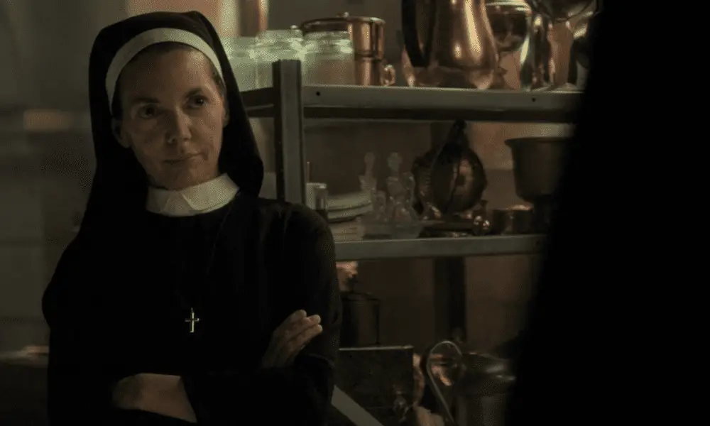 Sister Maggie stands with arms crossed and a skeptical look on her face. She is wearing her full nun habit. Shelves of church paraphenalia are in the background.