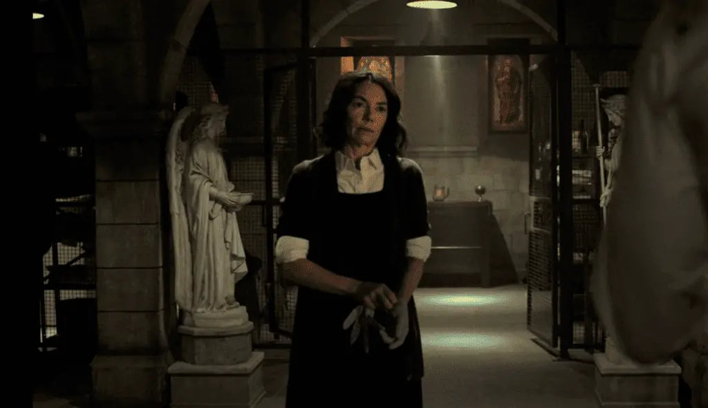 Sister Maggie stands in a church basement. She is wearing white shirtsleeves and a black dress, putting on blue gloves, looking exasperated. Angel statues and stained glass windows are in the background.
