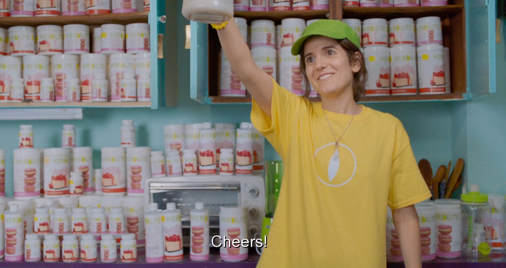 Tati, wearing a yellow t-shirt and green visor, raises a travel mug and smiles brightly, saying "Cheers!" In the background, the kitchen counter and all the cupboards are filled with jars and bottles of Hierbalite products.