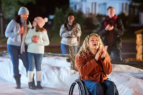 a blonde curly haired woman in a wheelchair wearing a rust orange jacket and jeans against backdrop of snow and people clapping