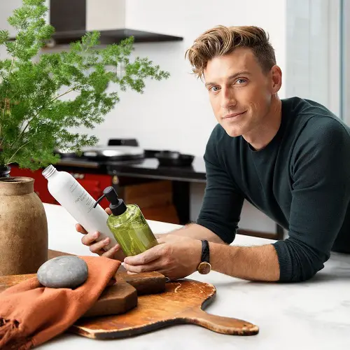 White mean wearing dark green long-sleeve shirt leaning over kitchen island with home products.