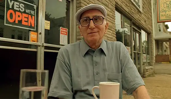 Junior Soprano sits outside a cafe, dressed in old-man slacks.