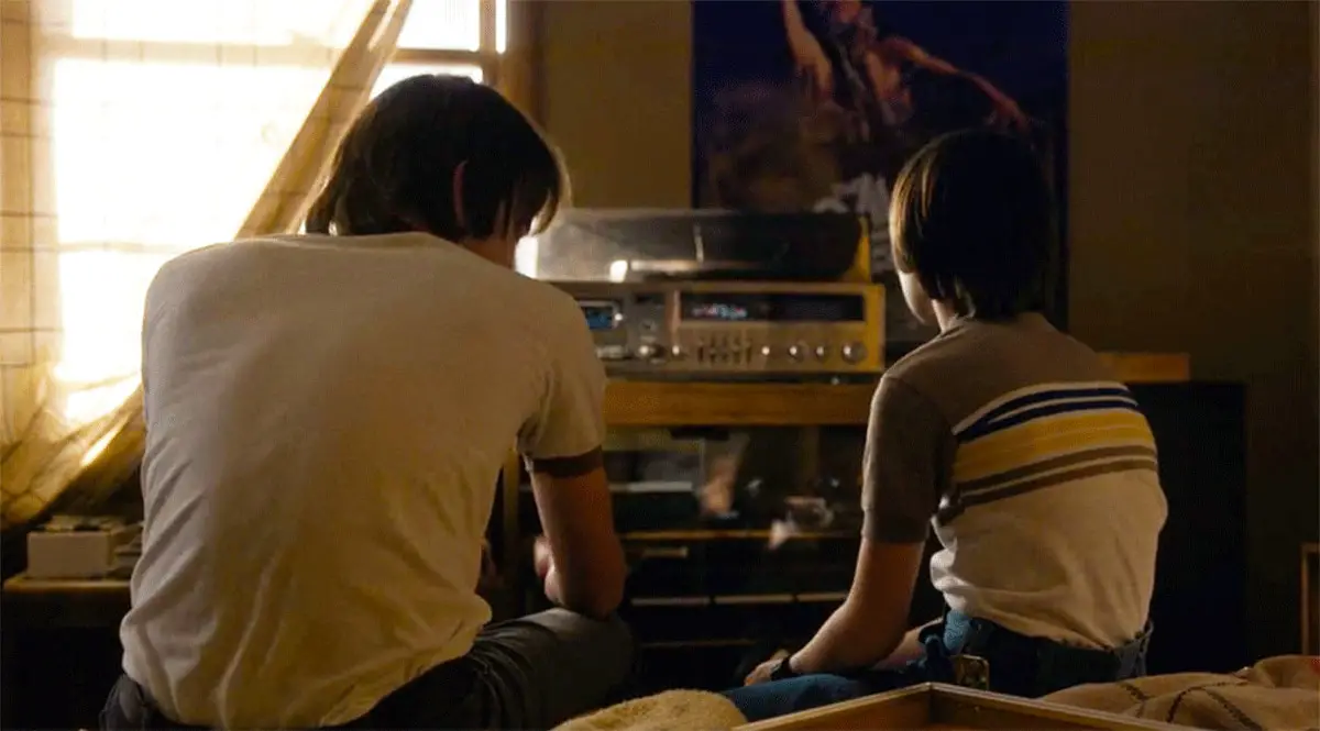 Jonathan (older brother) and Will (younger brother) from Stranger Things sit with their backs turned to the screen while they listen to a record playing. Conversation hints at Will's sexuality.