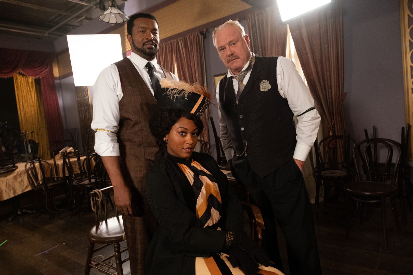 a Black man standing next to a white man with a Black woman sitting on a chair in front of them all dressed in 1900s clothing