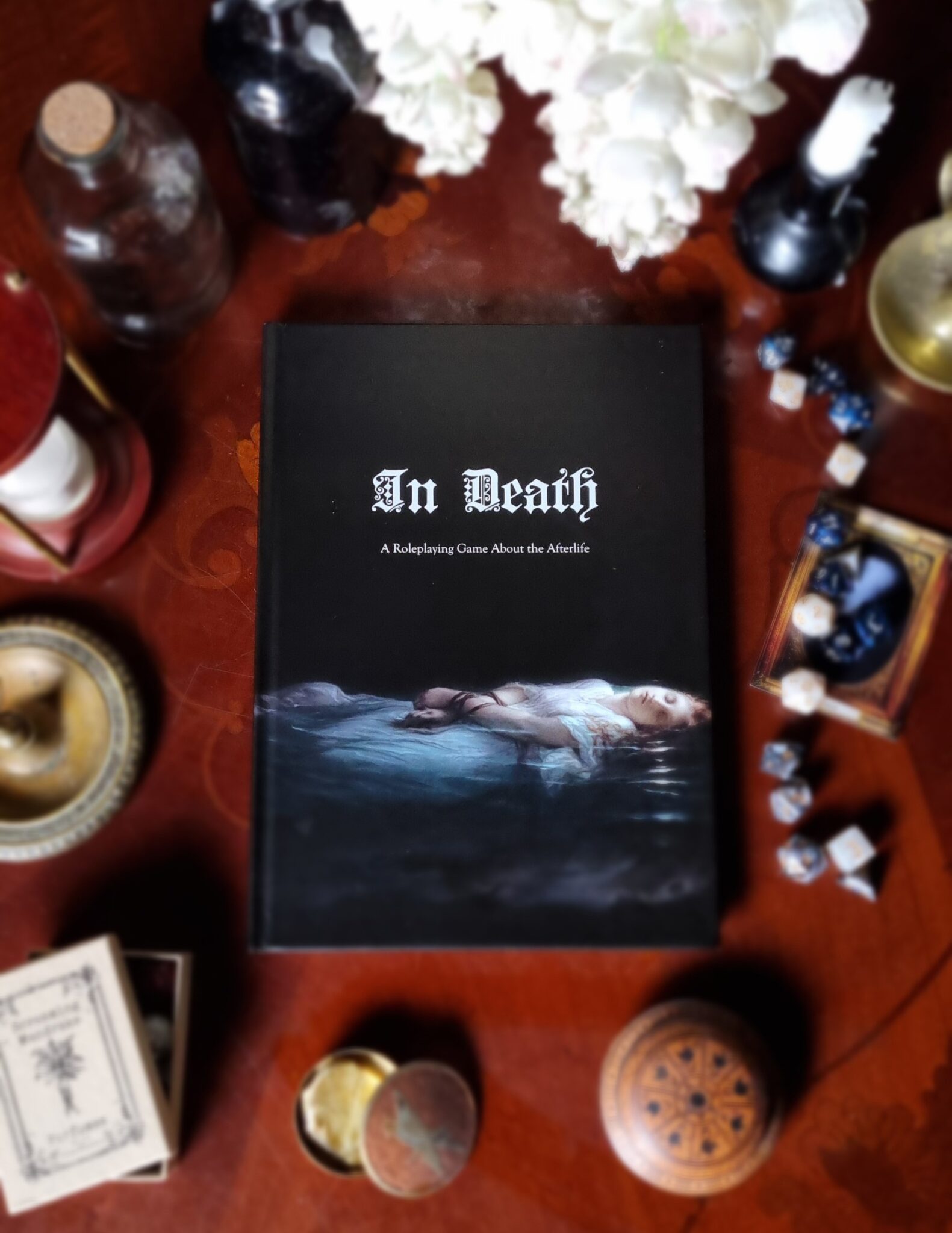 The TTRPG In Death, a black book with art of a woman in water and stylized white text. The background is blurred.